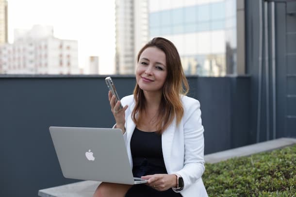 mujer joven sentada en el borde de un jardin, con su mano izquierda sostiene el notebook y con su mano derecha el celular